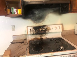 A kitchen stove with soot and smoke damage on the wall and countertop, indicating a fire incident.
