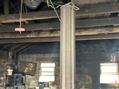A cluttered garage interior with exposed ceiling beams and a central metal support column, showing signs of fire damage and soot on the walls; tools, storage bins, and debris are scattered around, highlighting the need for fire damage mitigation and structural cleanup.