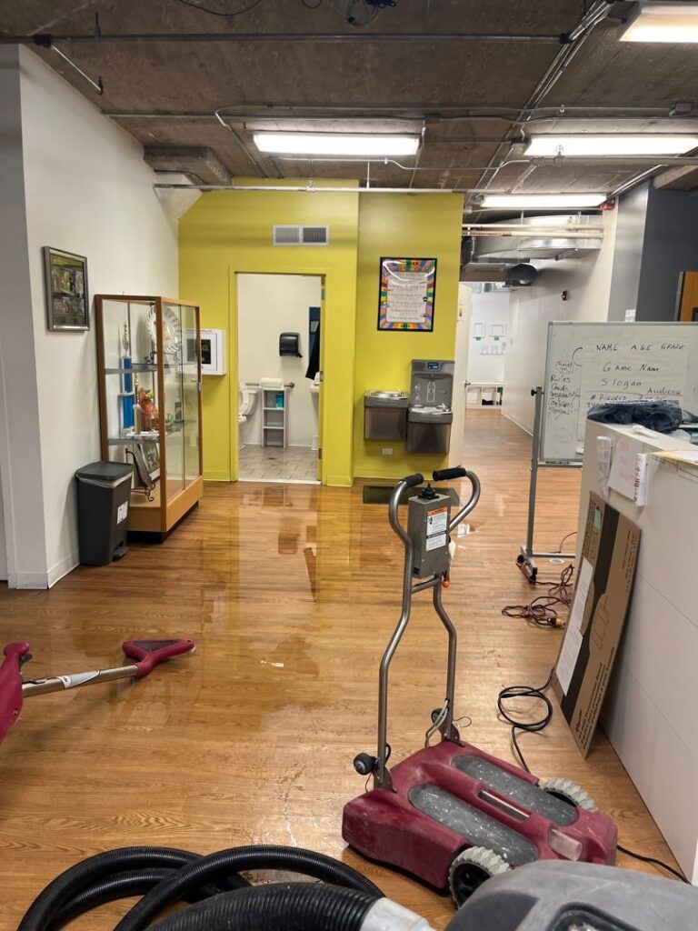 Office interior with hardwood floors flooded after a pipe burst, showing standing water, drying equipment, hoses, and cleanup tools in a commercial workspace.