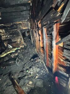 A photo showing the extensive damage inside a home after a severe house fire. The wooden wall studs are exposed and deeply charred, and the floor is completely covered in burnt debris and soot. A dark, smoke-damaged hallway is visible in the background.