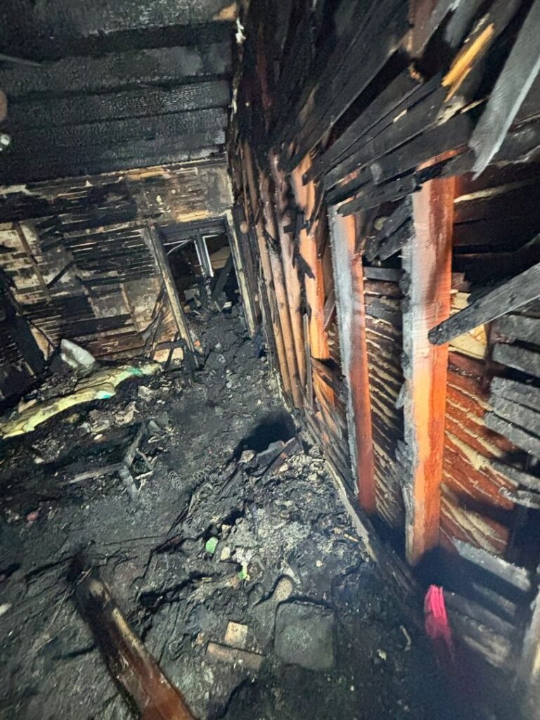 A photo showing the extensive damage inside a home after a severe house fire. The wooden wall studs are exposed and deeply charred, and the floor is completely covered in burnt debris and soot. A dark, smoke-damaged hallway is visible in the background.