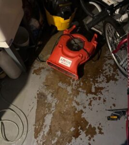 PuroClean red air mover drying standing water on a concrete floor caused by an appliance leak in a residential utility space.