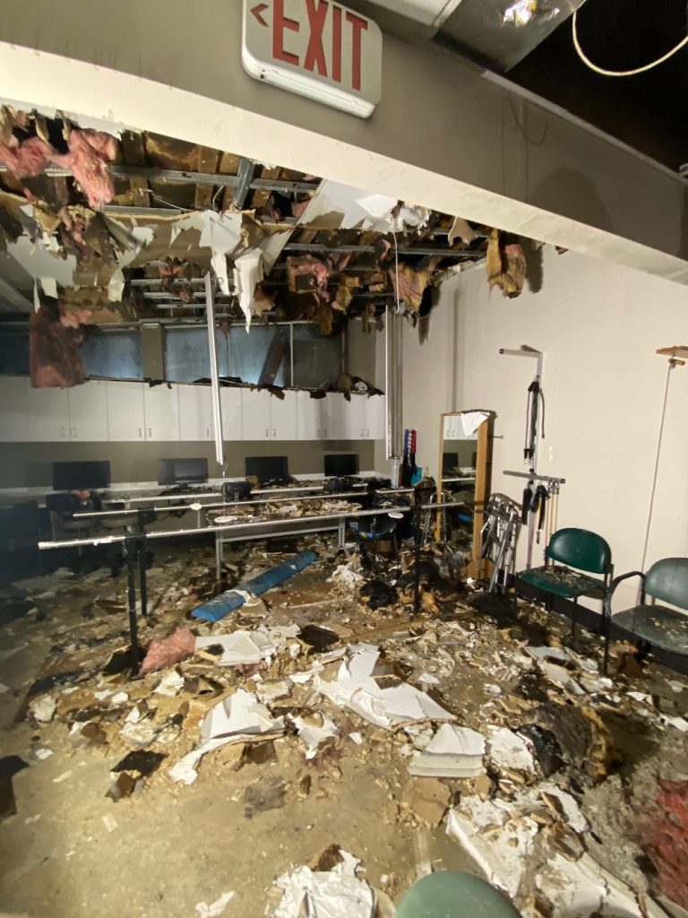 A severely damaged room features a collapsed ceiling, debris covering the floor, and an illuminated exit sign