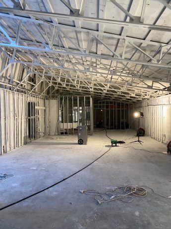 Spacious room under construction showing exposed metal stud framing, concrete floor, and equipment. TITLE