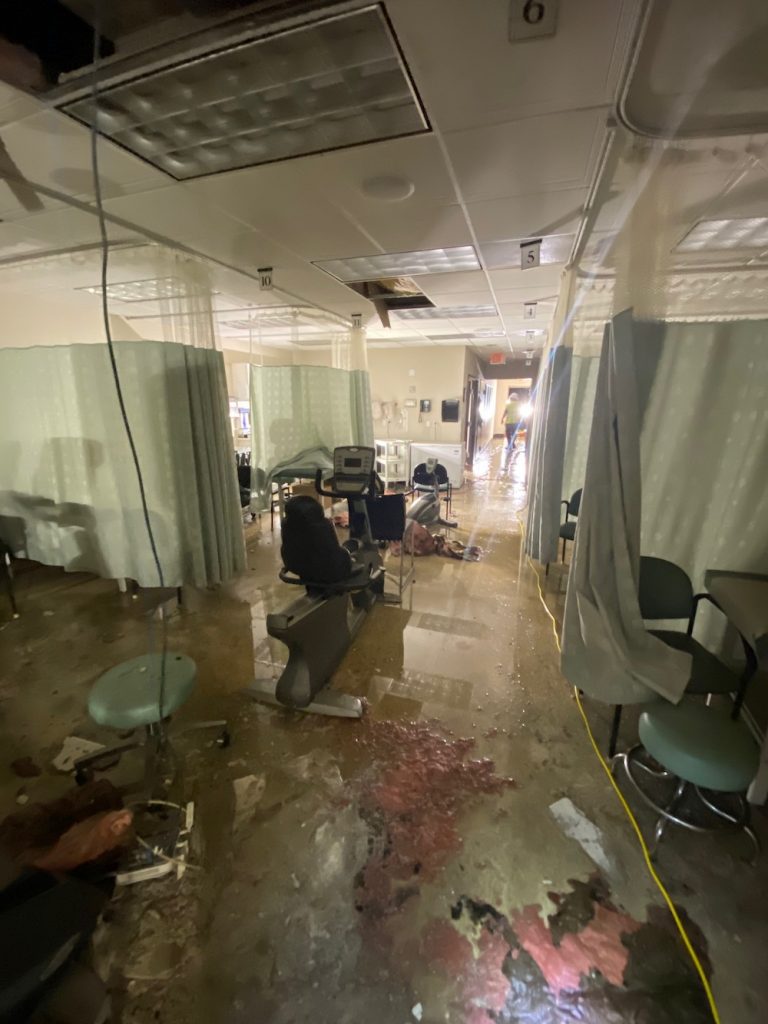 Severe water damage in a medical facility, with a flooded floor and collapsed ceiling tiles.