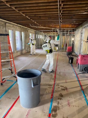 Two professionals in protective gear spray a large, unfinished room with exposed beams and metal studs.
