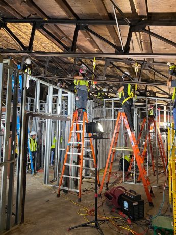Construction workers in safety gear are framing interior walls with metal studs on a job site.
