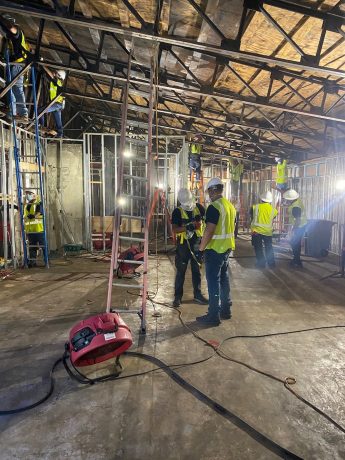 Construction workers in hard hats and safety vests install metal framing for interior walls and ceiling trusses.