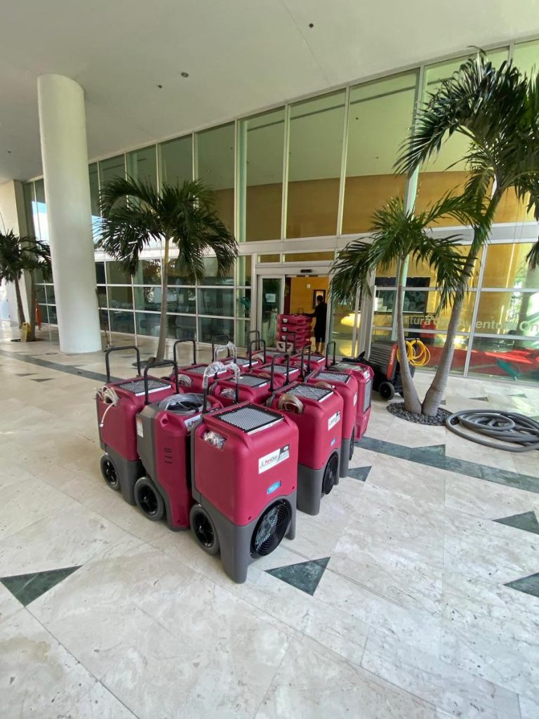 Several red industrial dehumidifiers are lined up on a marble floor in a modern building lobby.