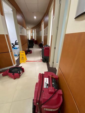 Red water damage restoration equipment and a caution sign are placed along a curved office hallway.