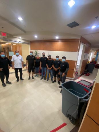 A team of masked service workers in black shirts stands together in a modern office hallway