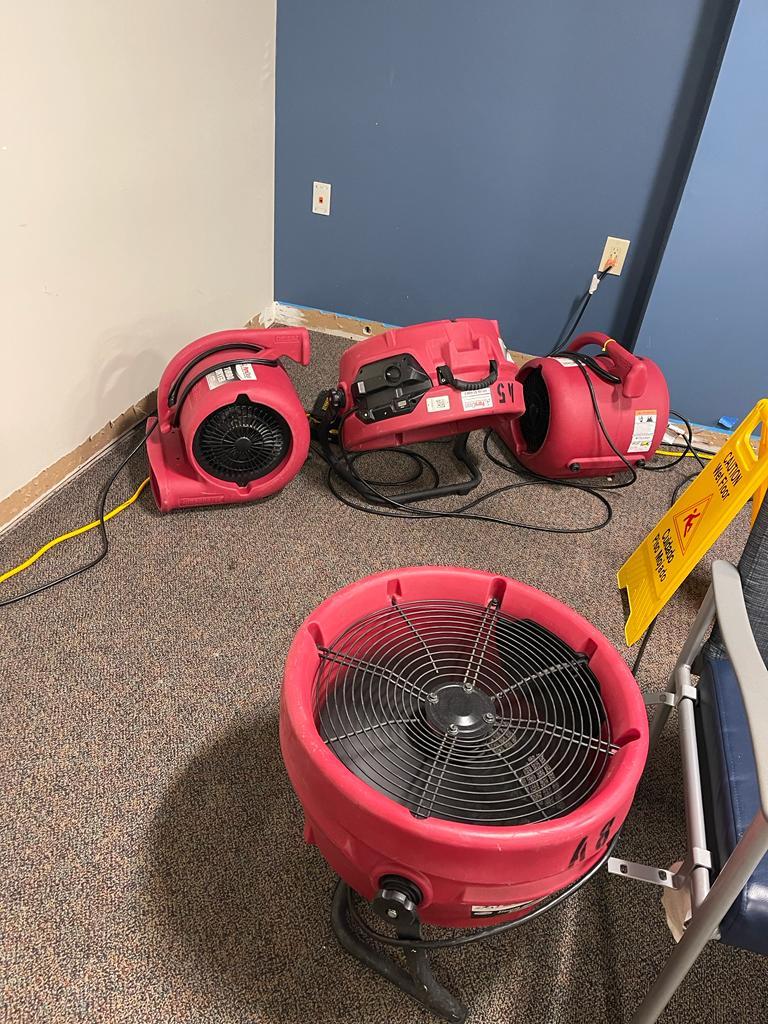 Red industrial fans dry a carpeted room with a blue wall and a wet floor sign.