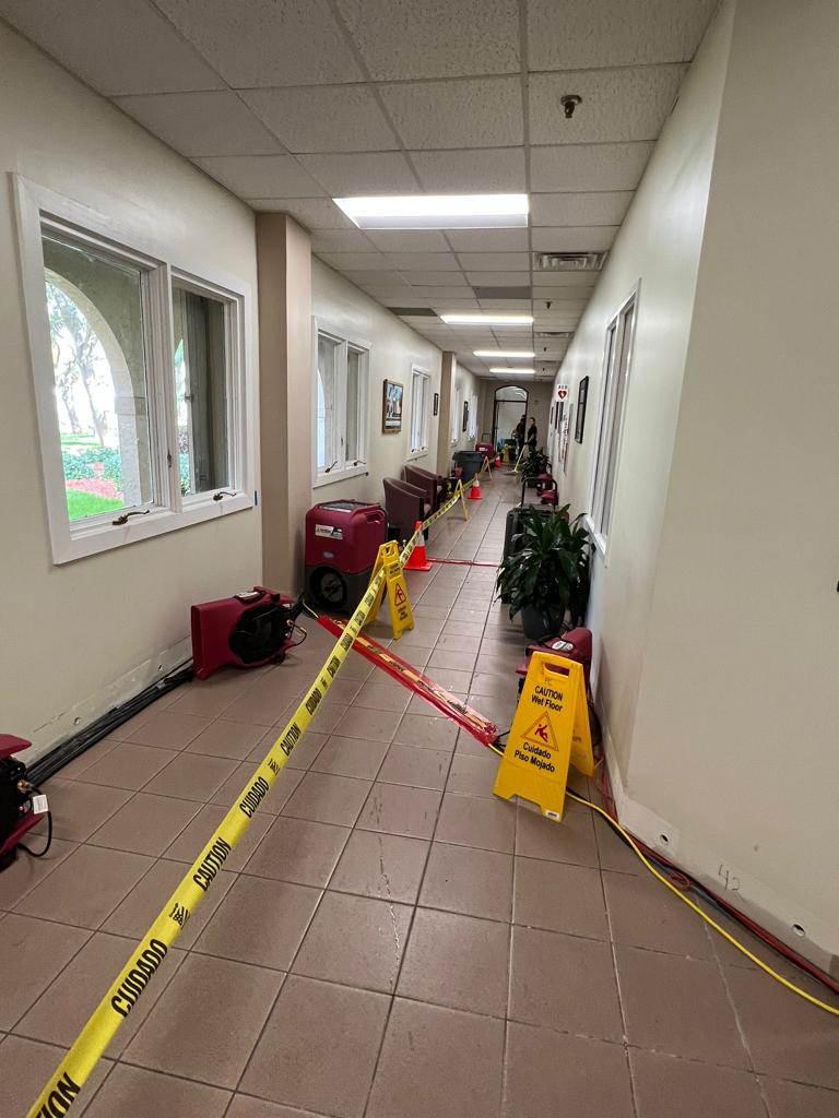A long hallway is blocked by caution tape and wet floor signs, with industrial fans for water