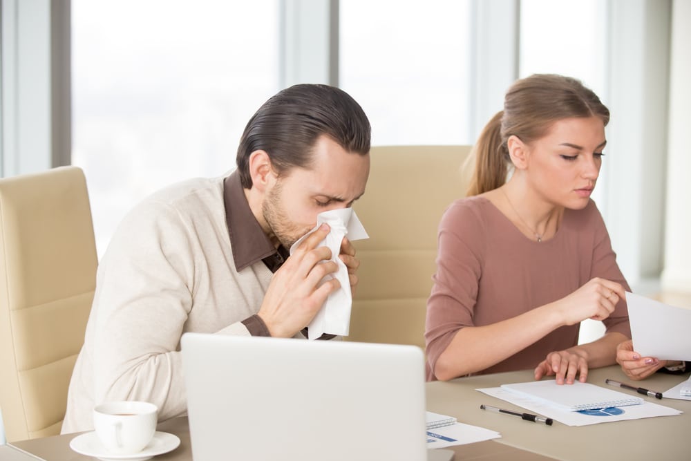 A man wipes his nose with a tissue while a woman reviews documents at a conference table in a modern office.