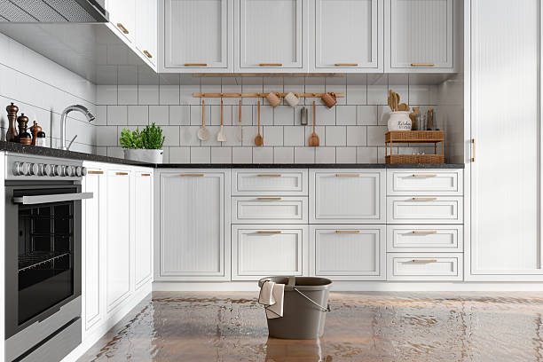 A modern kitchen with white cabinets is flooded with water, and a bucket sits on the wet floor