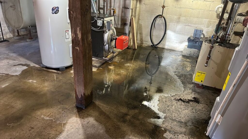 A utility basement floor is flooded with standing water around appliances and a wooden support beam