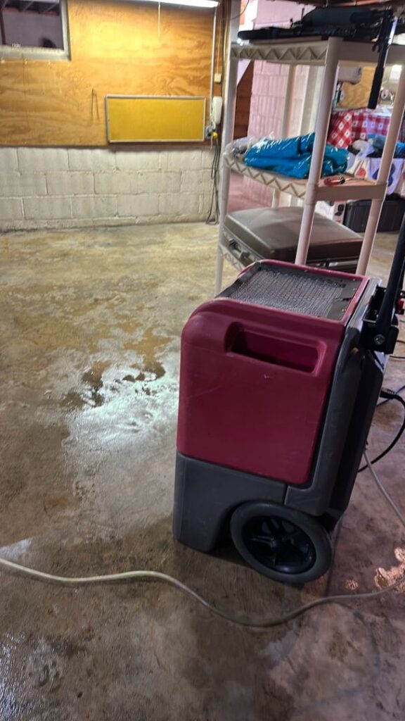 A red commercial dehumidifier sits on a wet, stained concrete basement floor with puddles.
