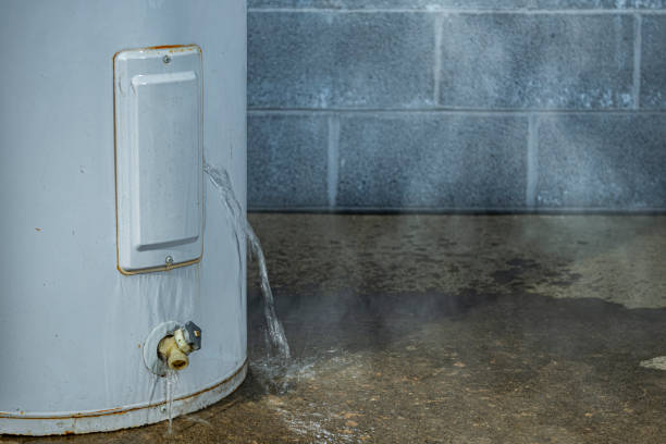 A white water heater leaks profusely onto a concrete floor, creating a puddle and steam.