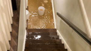 Murky floodwater fills a basement floor, with wet stairs leading down and buckets floating. this leads to Basement Flooding In Peoria