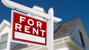 A red "FOR RENT" sign stands prominently on a white post in front of a blurred house.