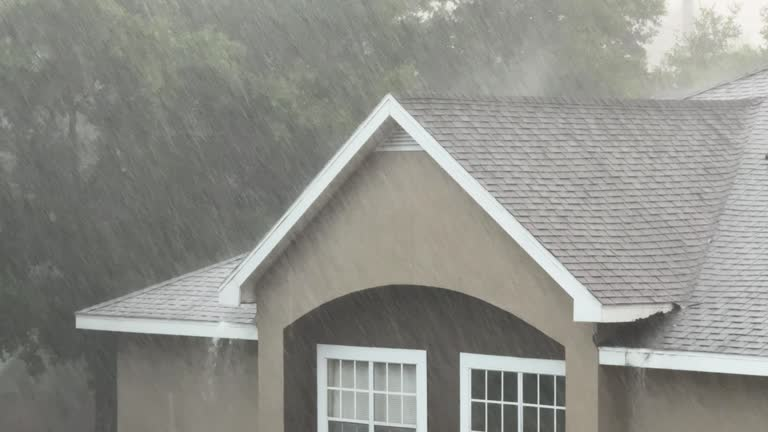 Streaks of heavy rain fall over a tan house with a gray roof and blurred trees. Professional Restoration