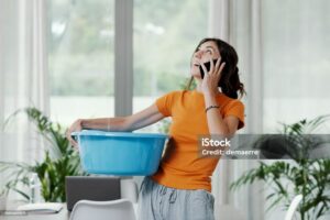 Concerned woman catches a ceiling leak in a blue basin while talking on her phone. TITLE