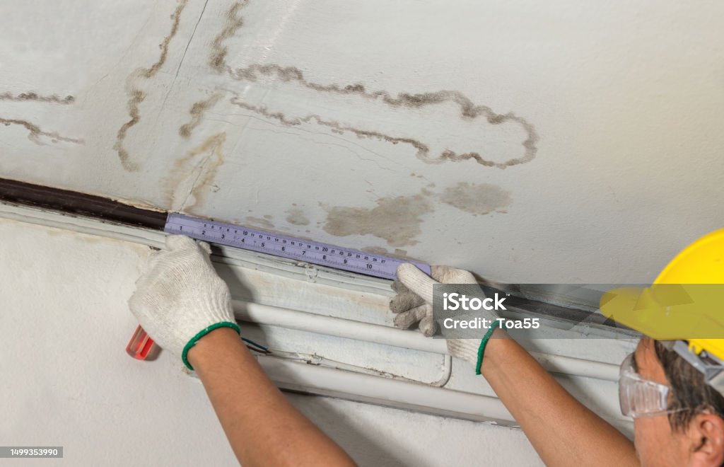 A worker in a hard hat measures water damage and mold on a ceiling. TITLE: