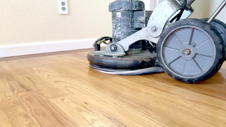 A close-up view of an industrial floor buffer in action, polishing a hardwood floor. The machine's rotating pad moves swiftly across the surface, creating a smooth, shiny finish. The worn equipment contrasts with the freshly polished wood, highlighting the effectiveness of the process. Ideal for themes related to floor care, professional cleaning, and home renovation. Perfect for content about hardwood floor maintenance, polishing techniques, and industrial cleaning equipment.