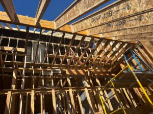 Extensive house framing and structural reconstruction and repairs due to fire damage, showing new wooden roof trusses, engineered I-joists, and scaffolding under a clear blue sky.