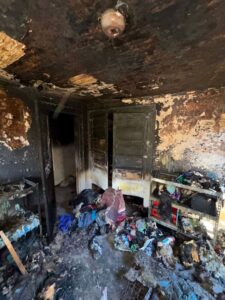 A photograph of a bedroom interior completely devastated by a recent house fire, with every surface blackened, walls peeling with charred paint, and piles of burnt clothing and debris covering the floor and shelves.