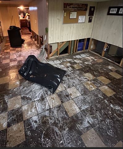 A basement room showing severe water damage and remediation efforts. The checkered tile floor is covered in a dirty, chalky white and brown water residue. Along the base of the wood-paneled walls, the lower section has been cut away and removed, exposing the internal wooden studs. A large black trash bag and a white plastic shovel rest in the center of the room.