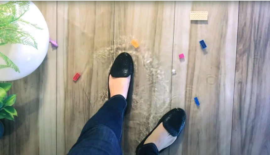 A person standing in water on a wooden floor, surrounded by colorful objects and a plant.