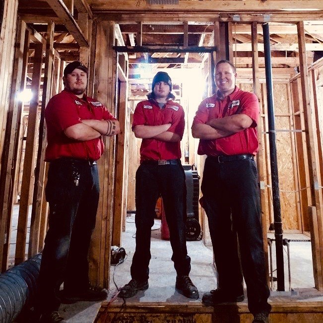 Three workers in red shirts stand confidently in a wooden-framed construction site, arms crossed.