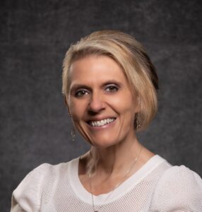 A smiling woman with short blonde hair and a white top looks directly at the viewer.