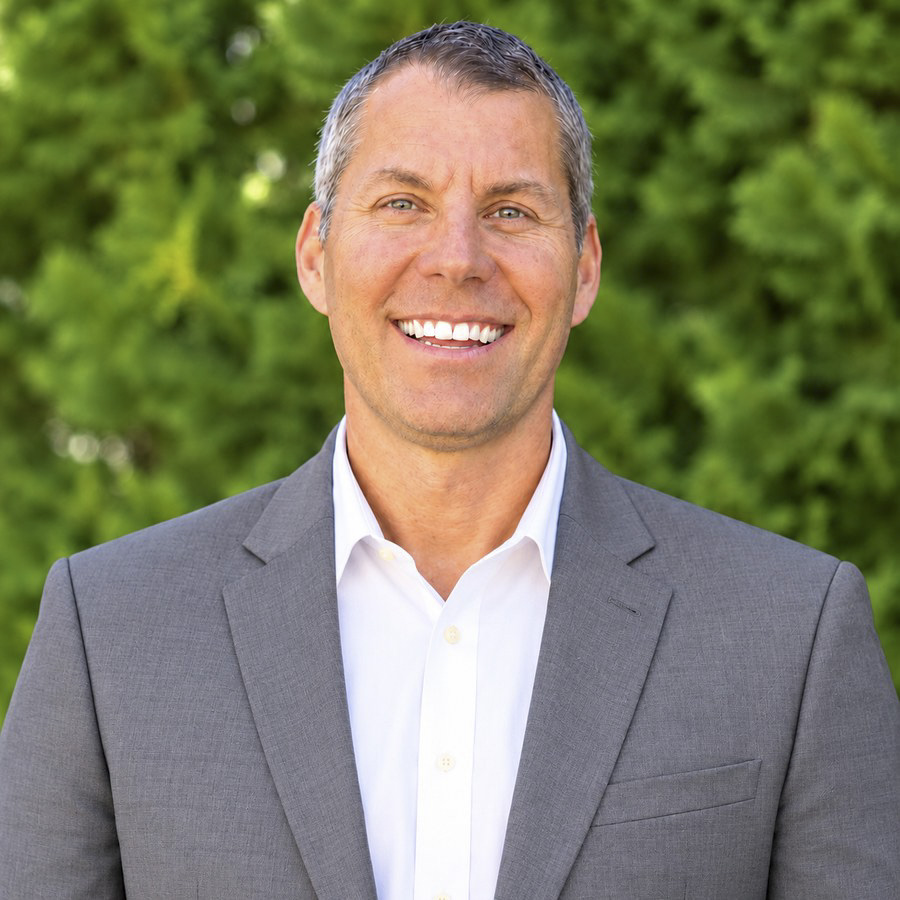 A friendly man in a gray suit smiles broadly against a blurred green background.