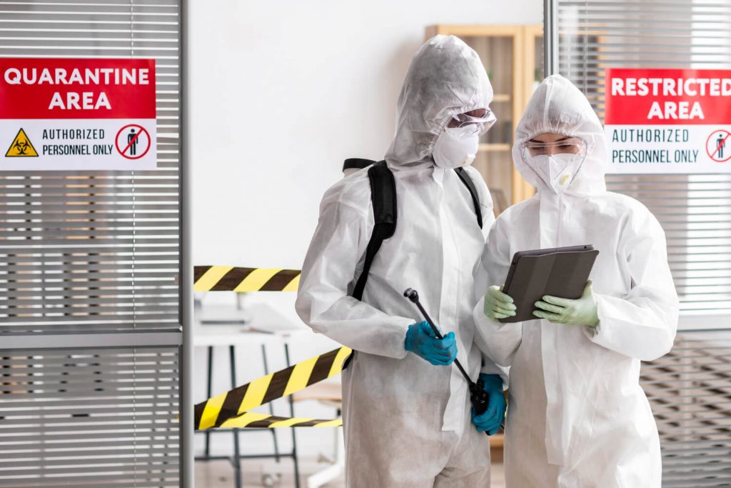 Two hazmat-suited personnel with a sprayer and tablet stand by a restricted quarantine area.