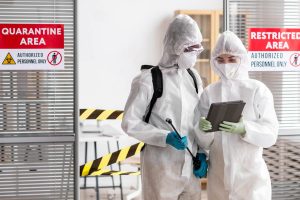 Two hazmat-suited personnel with a sprayer and tablet stand by a restricted quarantine area.