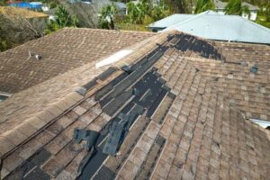 An aerial view of a residential brown shingle roof with extensive storm damage and missing shingles.