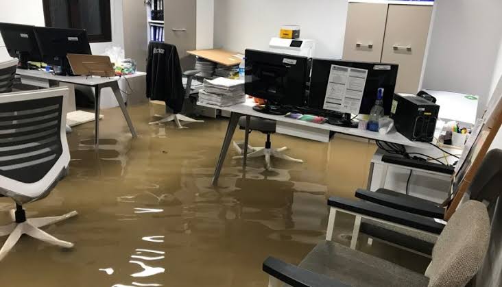 Muddy floodwater covers the floor of a modern office, submerging chair bases and lower desk legs.