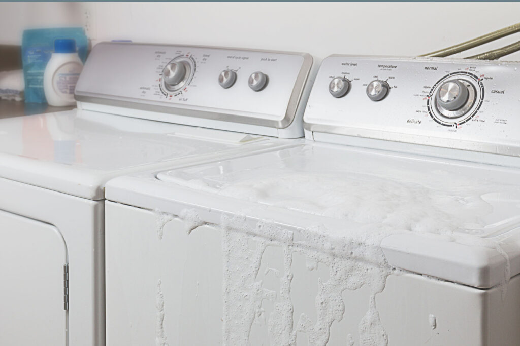 In a residential laundry room, a standard white washing machine (next to a standard white dryer) is overflowing with sudsy water. It's spewing out of the lid on the top of the machine—a common household property damage. 