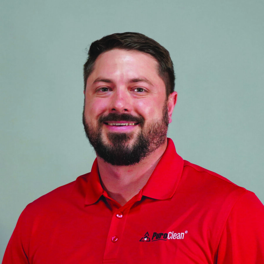 Smiling man in a red polo shirt with a logo, standing against a light gray background.