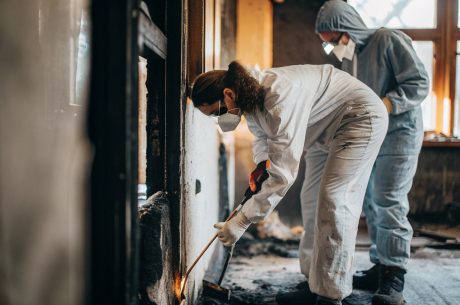 Two workers wearing protective suits, gloves, and face masks clean and inspect fire-damaged walls inside a building, removing debris and soot in a partially burned room.