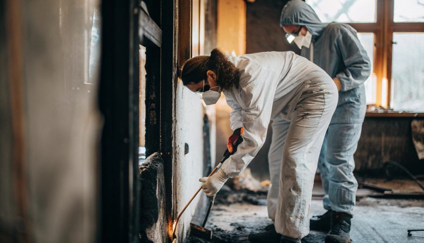 Two workers wearing protective suits, gloves, and face masks clean and inspect fire-damaged walls inside a building, removing debris and soot in a partially burned room.