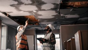 Two construction professionals wearing safety helmets inspect water damage on a ceiling inside a residential building