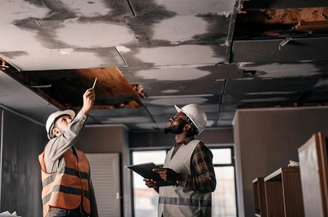 Two construction professionals wearing safety helmets inspect water damage on a ceiling inside a residential building
