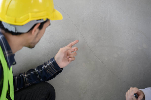 A construction worker in a yellow hard hat points to a diagonal crack in a concrete wall.