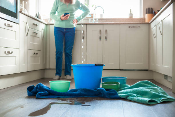 A senior woman looks worried about water damage as she deals with a water leak in the kitchen ceiling. Buckets and pots are on the floor to catch the flowing water, and towels are soaking up the puddles. The woman is defocused in the background, using her smart phone.