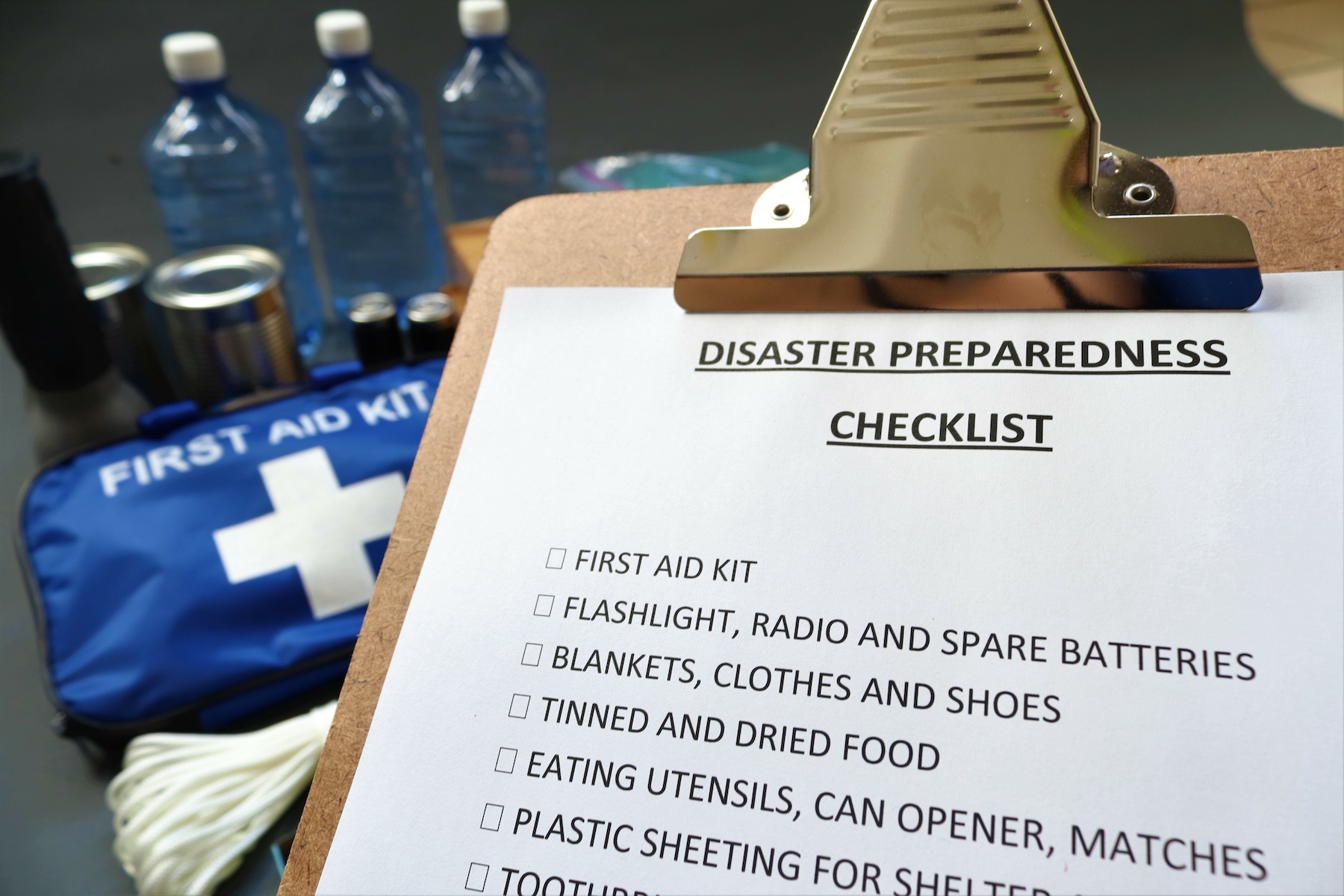 Disaster preparedness checklist on a clipboard with disaster relief items in the background.Such items would include a first aid kit,flashlight,tinned food,water,batteries and shelter.