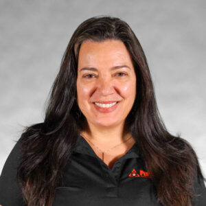 A woman with long dark hair smiles, wearing a black polo shirt with a logo, against a gray background.