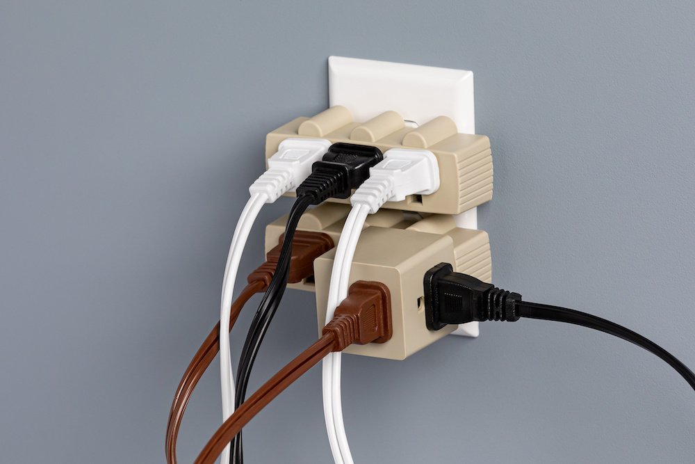 Several beige extension cords plugged into an outlet and overloaded with various plugs. Extension cord safety tips state that you should never overload your cords.
